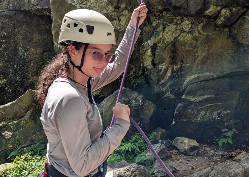 A young woman wearing a white climbing helmet and sunglasses is holding a purple rope. She is positioned in front of a rocky cliff face with some greenery at the base. She is wearing a long-sleeved grey shirt and has climbing gear around her waist. The lighting suggests it's daytime, and she appears to be preparing for or engaged in a climbing activity.